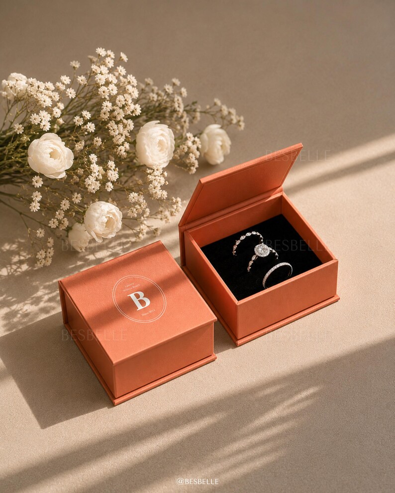 May include: Two square terracotta-colored jewelry boxes with hinged lids. One box is closed, displaying a white circular logo with the letter B. The other box is open, revealing a black velvet interior holding three silver-toned rings with stones.