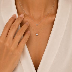 以下が含まれることがあります： 2本の繊細なチェーンを重ねたネックレスセット。短いチェーンはシルバーカラーで小さな丸いソリティアストーンが付いており、長いチェーンはゴールドカラーで大きめの丸いソリティアストーンが付いています。BESBELLEという文字が見えます。