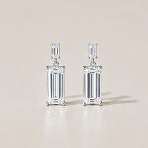 May include: A pair of elegant silver earrings with rectangular, clear gemstones. Each earring features a smaller rectangular stone at the top, connected to a larger, elongated stone below. The earrings are set against a neutral background.