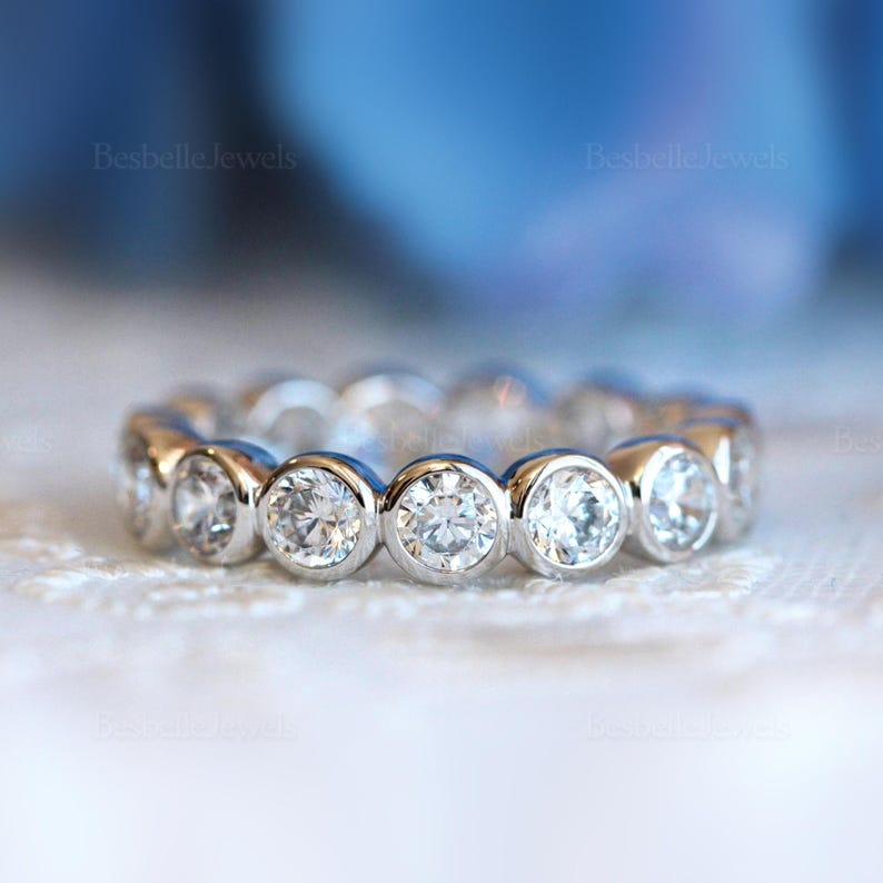 May include: A silver eternity ring with round, clear gemstones set in individual bezels. The ring is displayed on a white, textured surface with a soft blue background. The text "Besbelle Jewels" is visible.