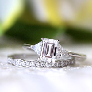 May include: A silver engagement ring set with a large emerald cut center stone and two smaller triangular stones on either side. The ring is paired with a delicate silver band with a scalloped edge.