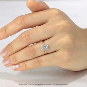 May include: A gold ring with a cluster of small, clear stones surrounding a larger, oval-shaped stone. The ring is on a person's finger, with the hand in the frame. The ring is set against a neutral background. The text "Besbelle Jewels" is visible.