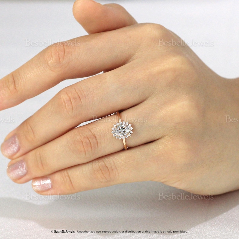 May include: A gold ring with a cluster of small, clear stones surrounding a larger, oval-shaped stone. The ring is on a person's finger, with the hand in the frame. The ring is set against a neutral background. The text "Besbelle Jewels" is visible.