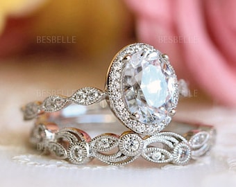 May include: A silver-toned engagement ring set featuring an oval-cut center stone surrounded by a halo of small round stones. The band and the matching contoured wedding band are decorated with intricate metalwork and embedded round stones.