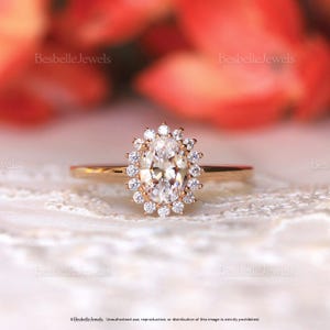 May include: A gold ring with a large oval-cut diamond center stone, surrounded by a halo of smaller diamonds. The ring is set on a delicate band. The background is blurred, with a soft, floral-like pattern. The ring is a piece of jewelry.