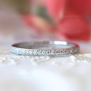 May include: A delicate, silver-toned ring with a continuous row of small, clear gemstones. The ring's band is thin and features a detailed, textured edge. The background is soft and out of focus, with the text "Besbelle Jewels" visible.