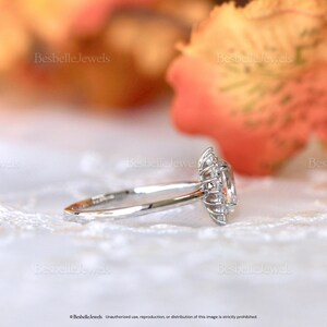 May include: A silver engagement ring with a large, oval-shaped center stone surrounded by smaller accent stones. The ring is set against a soft, blurred background with the text "Besbelle Jewels".