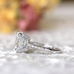 May include: A silver ring with a large, square-cut, clear gemstone at the center, surrounded by smaller stones. The band is also adorned with small, sparkling stones. The ring is set against a soft, blurred background.