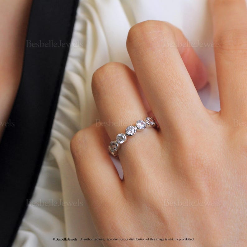 May include: A silver ring with a series of round, clear gemstones set in a row. The ring is on a person's finger. The image has the text "Besbelle Jewels".