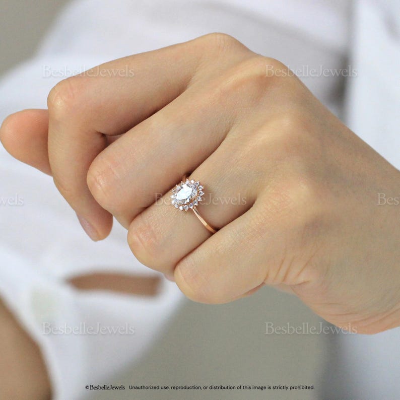May include: A rose gold ring featuring a central oval-cut gemstone surrounded by a halo of smaller stones. The ring is on a finger, showcasing its design and sparkle. The ring's band is thin and delicate, complementing the overall elegance.