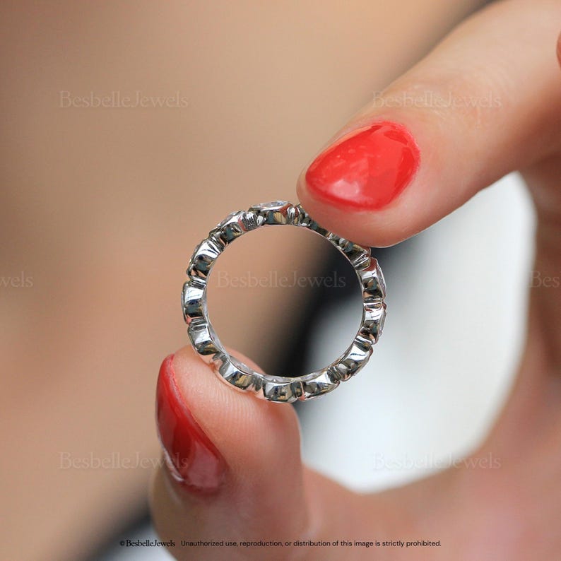 May include: A silver eternity ring with a repeating pattern of small, clear gemstones. The ring is held between two fingers with red nail polish. The ring is a delicate piece of jewelry.