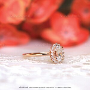 May include: A rose gold engagement ring with a large oval-cut center stone surrounded by a halo of smaller diamonds. The ring is set on a delicate band, and the background is blurred with red and white colors.