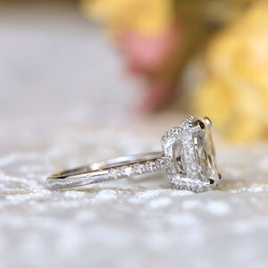 May include: A silver engagement ring with a large, rectangular, clear gemstone. The ring features a halo of smaller stones around the main stone and along the band. The ring is set on a white, textured surface.