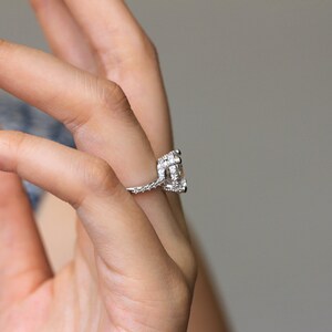 May include: A silver engagement ring with a large, clear, rectangular-cut center stone. The band and setting are encrusted with smaller, sparkling stones. The ring is held up by a hand, showcasing the intricate details of the jewelry.