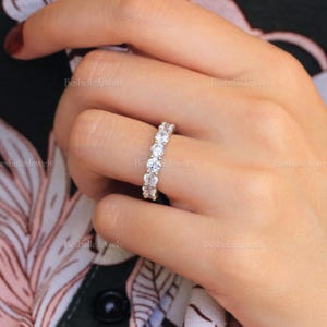 May include: A close-up of a diamond eternity ring on a finger. The ring features a row of round-cut diamonds set in a band. The ring is set against a background of a floral pattern.
