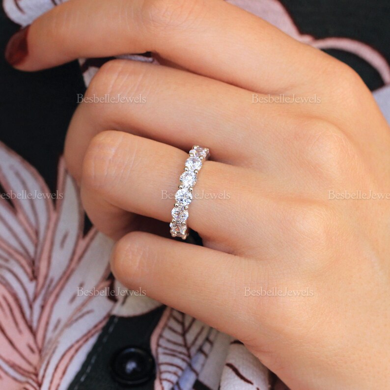 May include: A close-up of a diamond eternity ring on a finger. The ring features a row of round-cut diamonds set in a band. The ring is set against a background of a floral pattern.