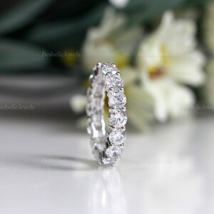 May include: A close-up of a silver eternity ring with multiple round, clear gemstones. The ring is set against a blurred background of white flowers and the text "BesbelleJewels". The ring is a symbol of commitment and love.