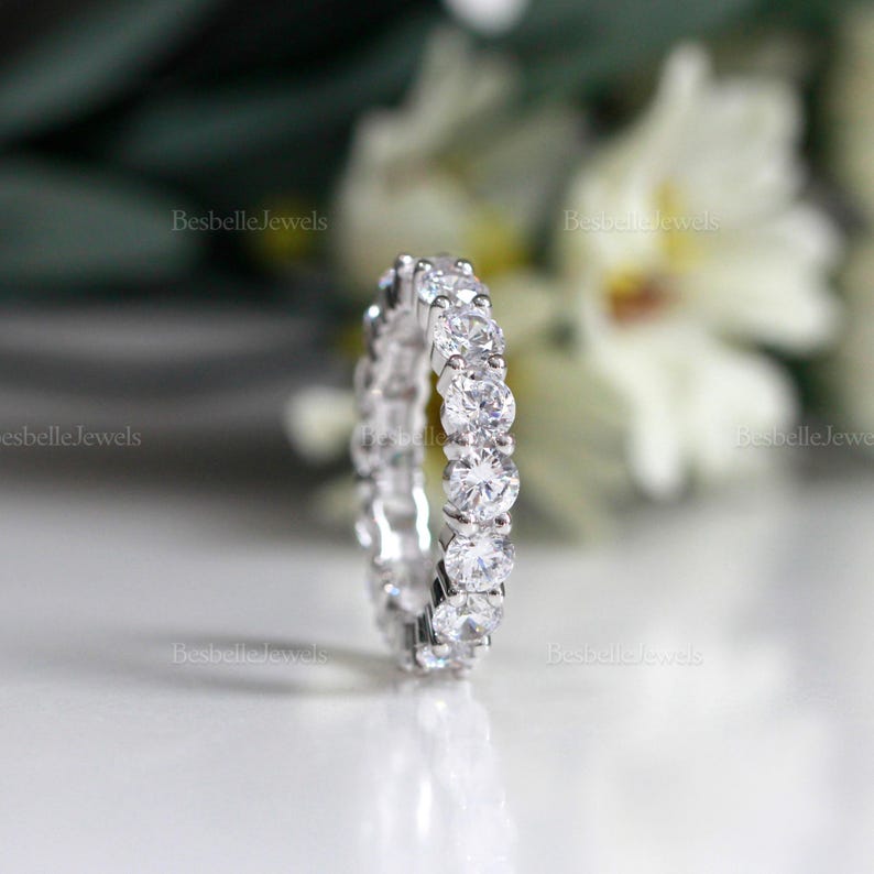 May include: A close-up of a silver eternity ring with multiple round, clear gemstones. The ring is set against a blurred background of white flowers and the text "BesbelleJewels". The ring is a symbol of commitment and love.