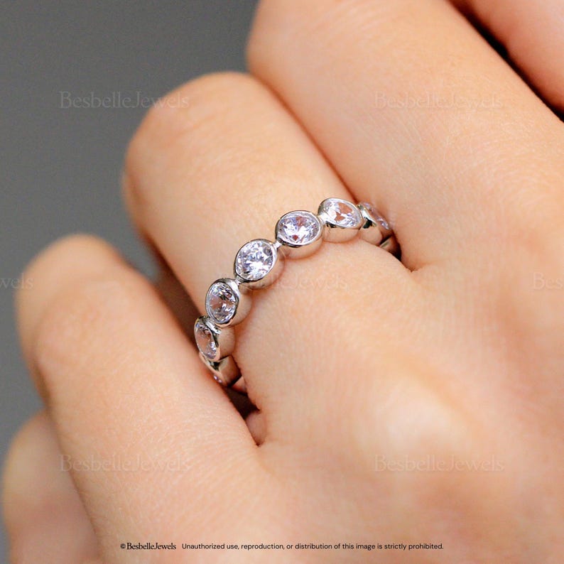 May include: A silver ring with a series of clear, round-cut gemstones set in a bezel style. The ring is shown on a person's finger, with the gemstones catching the light. The ring is a piece of jewelry.
