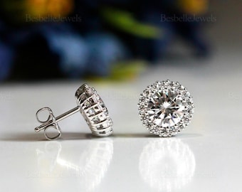 May include: A pair of silver stud earrings with a round, clear center stone surrounded by smaller, clear stones. The earrings are set on a white surface with a blurred background.
