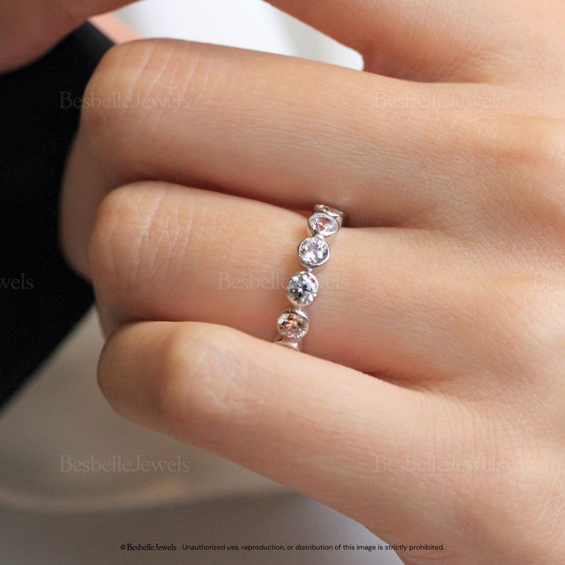 May include: A silver ring with five round, clear gemstones. The ring is set with bezel settings, and the gemstones are evenly spaced around the band. The ring is on a finger.