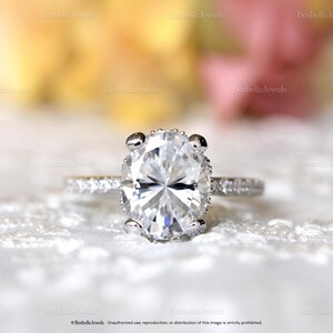 May include: A close-up of a diamond ring with an oval-cut center stone. The ring has a delicate band with smaller stones and four prongs holding the main diamond. The background is blurred, with soft colors.
