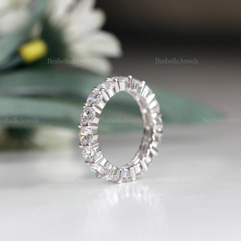 May include: A silver eternity ring with multiple clear, round-cut gemstones. The ring is set with a continuous line of stones, creating a sparkling effect. The ring is displayed on a white surface with a blurred background.