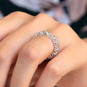 May include: A silver eternity ring with multiple clear, round-cut gemstones. The ring is set in a silver band and is displayed on a person's finger. The ring is from BesbelleJewels.