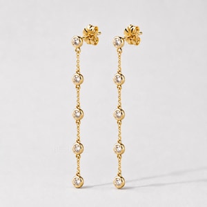 May include: A pair of gold dangle earrings with a delicate chain design. Each earring features a small, round, clear gemstone at the top, followed by four more gemstones suspended along the chain. The earrings have a polished finish and a classic, elegant style.