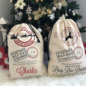 May include: Two white canvas Santa sacks with black drawstrings. The sacks have red and black printed designs with the text "Do Not Open Before December 25th" and "Special Overnight Delivery For:". One sack is personalised with the name "Charles" and the other with the name "Lucy Rae Duncan".