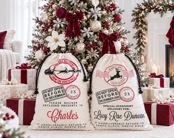 May include: Two off-white canvas drawstring gift sacks with black rope ties. Each sack features red and black printed graphics including a North Pole postmark, a reindeer silhouette, and the text 'Do Not Open Before 25 December'. The names 'Charles' and 'Lucy Rae Duncan' are printed in red and black script.