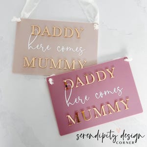 May include: Two signs with the text "Daddy here comes Mummy" in gold letters. The signs are made of acrylic and have a pink and beige colour scheme.