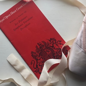 Puede incluir: Un programa rojo para una actuación de Les Patineurs, Variaciones Sinfónicas y Perséfone en el Royal Opera House Covent Garden el miércoles 29 de noviembre de 1977. Un zapato de ballet blanco con una cinta blanca está en primer plano.
