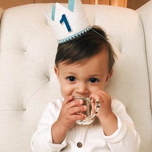 May include: A baby wearing a white and blue crown with the number "1" on it, sitting in a white chair and holding a small cup.