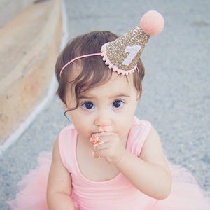 May include: A young child wearing a pink tutu and a gold glitter birthday hat with the number "1" on it. The child is eating cake and has frosting on their face.