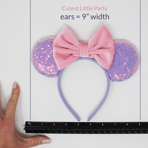 May include: A headband featuring two circular ears covered in iridescent purple sequins and a large, central pink velvet bow. The text Cutest Little Party ears = 9" width is visible above the headband, which is positioned next to a black ruler.
