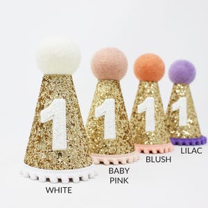 May include: Five gold glitter party hats with white pom pom trim and a white number one on the front. The hats are in different colors: white, baby pink, blush, and lilac.