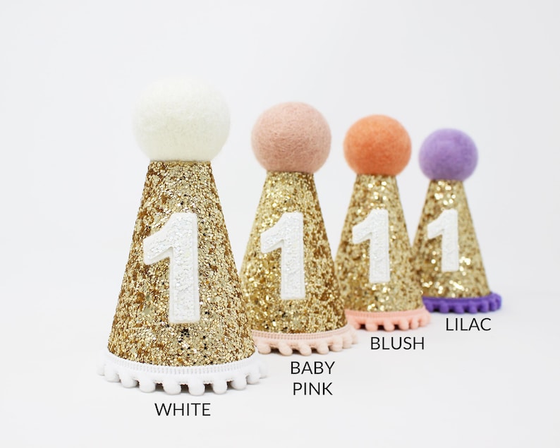 May include: Five gold glitter party hats with white pom pom trim and a white number one on the front. The hats are in different colors: white, baby pink, blush, and lilac.
