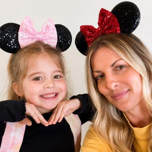 May include: Two headbands featuring round black sequined ears. One headband has a light pink sequined bow, and the other headband has a bright red sequined bow. Both headbands are constructed with a thin, flexible band to secure the ears in place.