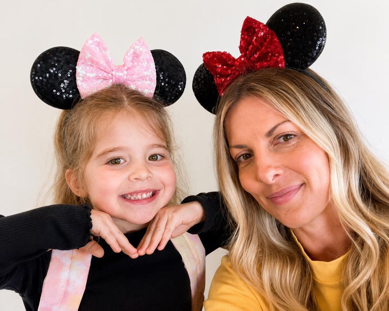 May include: Two headbands featuring round black sequined ears. One headband has a light pink sequined bow, and the other headband has a bright red sequined bow. Both headbands are constructed with a thin, flexible band to secure the ears in place.