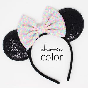 May include: A black headband featuring two round ears covered in black sequins and a large, central bow covered in iridescent, multi-colored sequins. The center of the headband displays the black text choose color in a simple, sans-serif font.