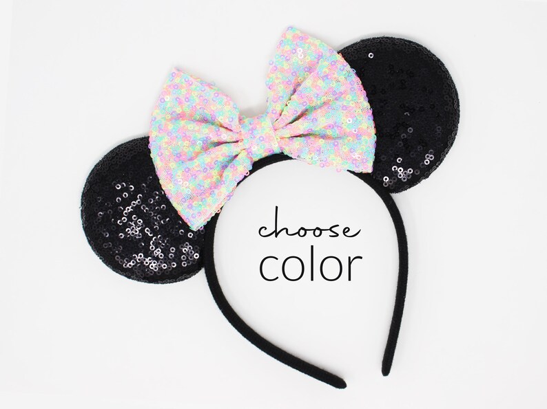 May include: A black headband featuring two round ears covered in black sequins and a large, central bow covered in iridescent, multi-colored sequins. The center of the headband displays the black text choose color in a simple, sans-serif font.