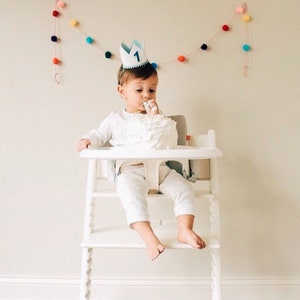 May include: A baby wearing a blue and white crown with the number "1" on it, sitting in a white high chair and eating cake. A colorful pom-pom garland hangs on the wall behind the baby.