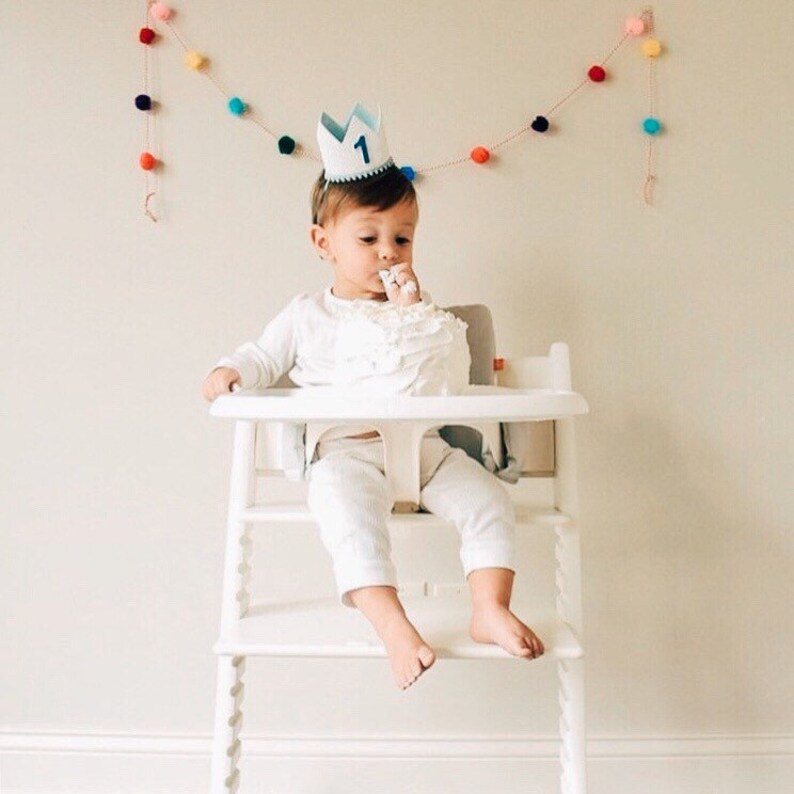 May include: A baby wearing a blue and white crown with the number "1" on it, sitting in a white high chair and eating cake. A colorful pom-pom garland hangs on the wall behind the baby.