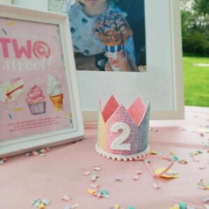 May include: A small, crown-shaped party hat featuring a glittery rainbow gradient finish, a white felt number two centered on the front, and a delicate white pom-pom trim along the bottom edge, accompanied by a thin pink elastic chin strap.