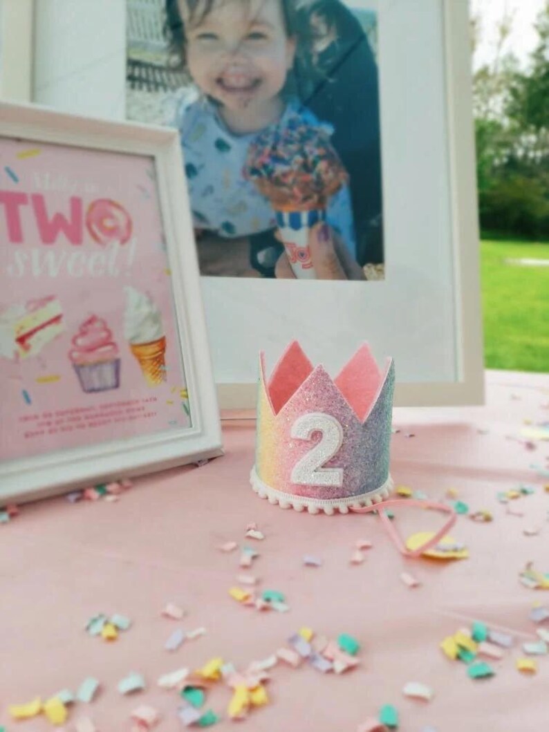 May include: A small, crown-shaped party hat featuring a glittery rainbow gradient finish, a white felt number two centered on the front, and a delicate white pom-pom trim along the bottom edge, accompanied by a thin pink elastic chin strap.