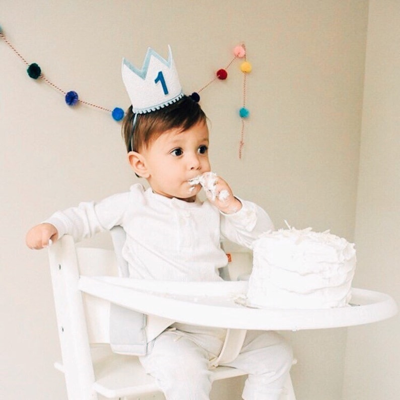 May include: A baby wearing a blue and white crown with the number "1" on it, sitting in a white high chair and eating cake.