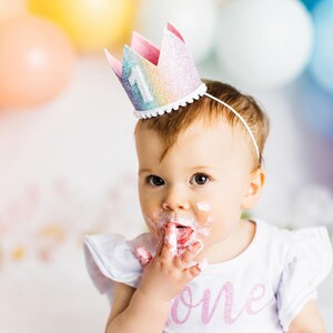 May include: A glittery rainbow-colored crown headband featuring a white number one on the front, worn by a child with white frosting on their face. The crown has a white pom-pom trim along the base and is attached to a thin white headband.
