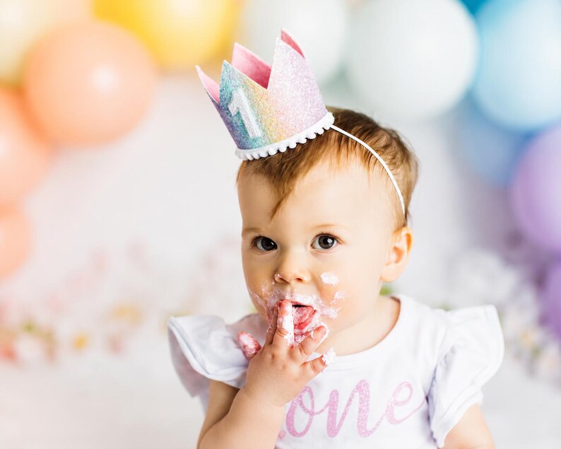 May include: A glittery rainbow-colored crown headband featuring a white number one on the front, worn by a child with white frosting on their face. The crown has a white pom-pom trim along the base and is attached to a thin white headband.