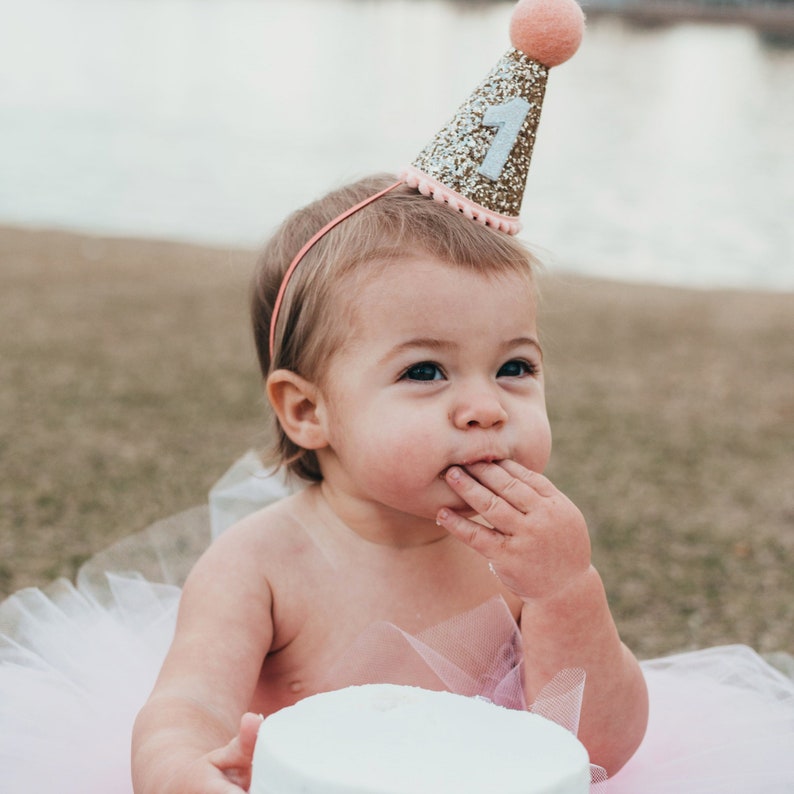 May include: A baby girl wearing a pink and gold glitter birthday hat with the number "1" on it. She is wearing a pink tutu and is eating cake.